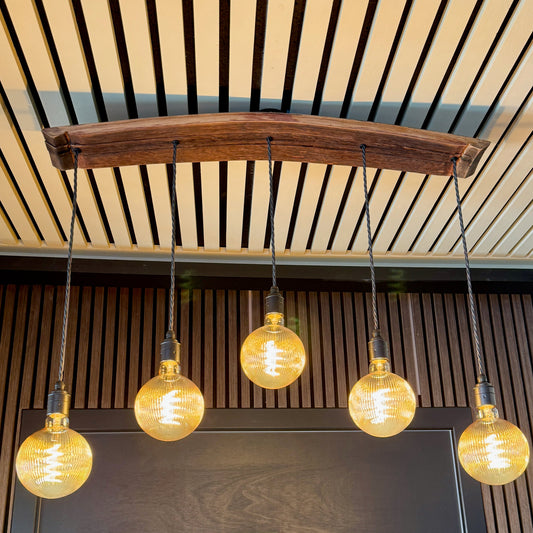 A rustic industrial-style pendant light fixture with a curved dark wood beam and five hanging warm white Edison-style bulbs, mounted against a slatted wood wall and ceiling.