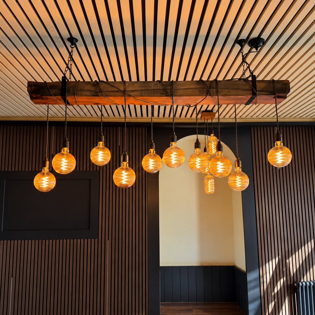 A rustic industrial chandelier with a thick wooden beam and multiple hanging Edison-style bulbs, installed in a modern interior with dark wood slat walls and a slatted ceiling.